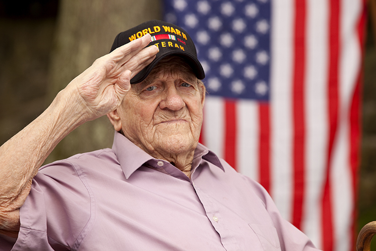 Older man wearing cap that reads "World War II Veteran" salutes with United States flag in background