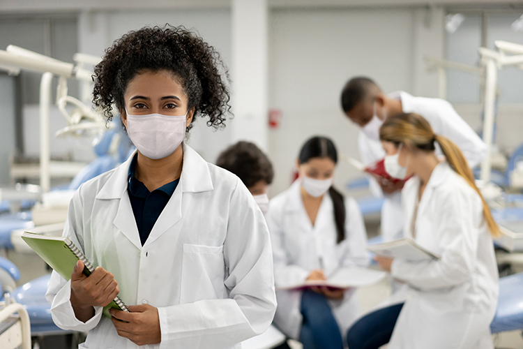 woman wearing surgical mask stands in foreground holding notebook while masked colleagues compare notes in background