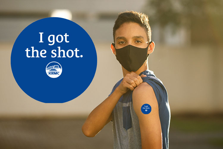 An adolescent boy wearing a face mask stands with his left shoulder bared and a sticker on his arm reads "I got the shot."