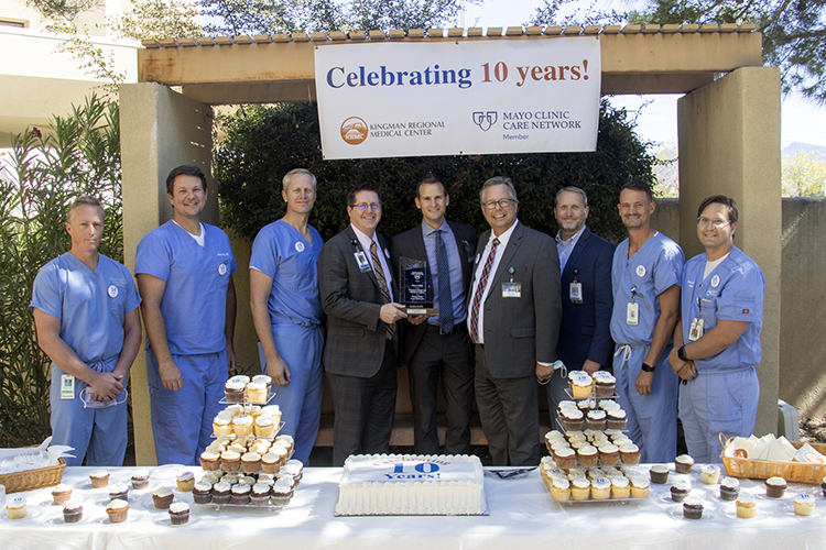 a group of nine white men including physicians and executives stand before a banner that reads "celebrating 10 years!" with a table of cakes in the foreground