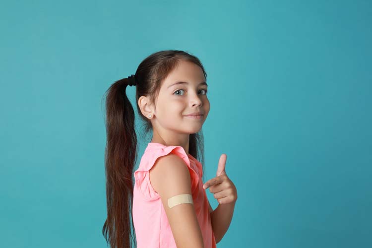 a child wearing a pink shirt and long pigtails looks into the camera, pointing with her left hand to a bandage on her right arm