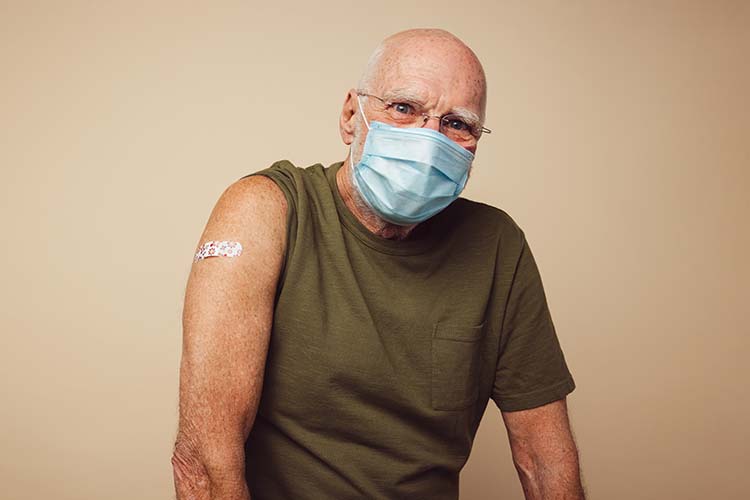 an older man in a mask and glasses sits with his right sleeve rolled up and a bandage on his arm