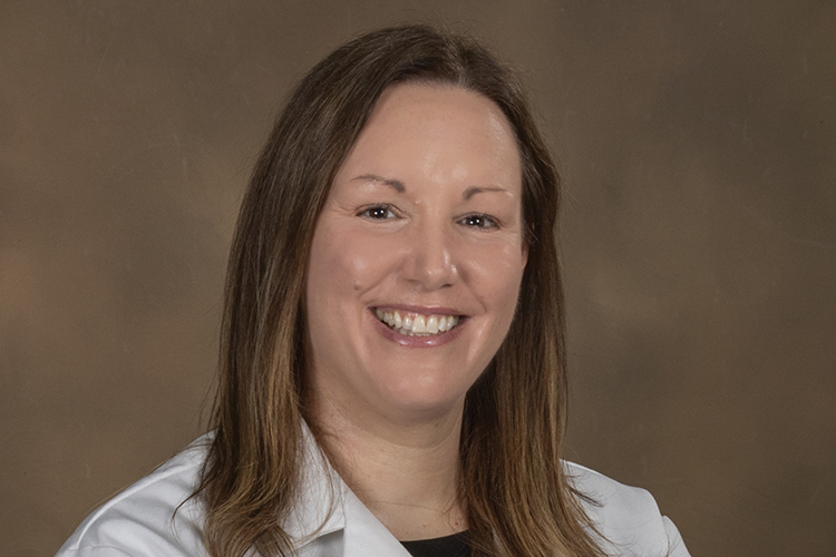 Dr. Joy Dunn smiles in a white lab coat before a tan background