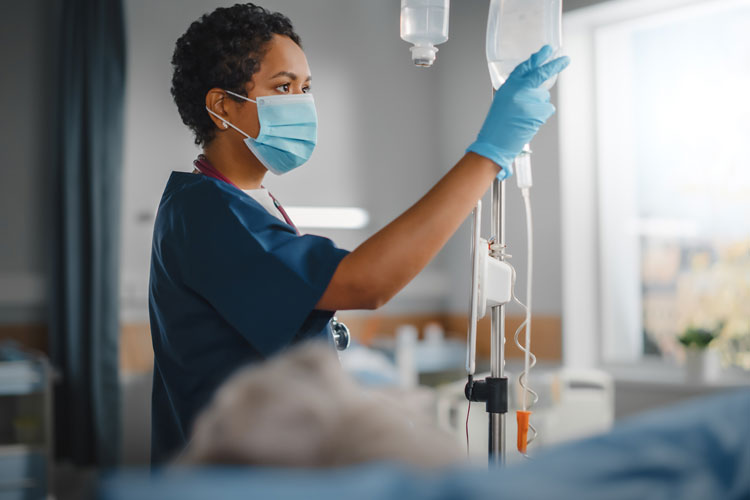 a medical professional in scrubs with a surgical mask and gloves touches an IV bag in a hospital room