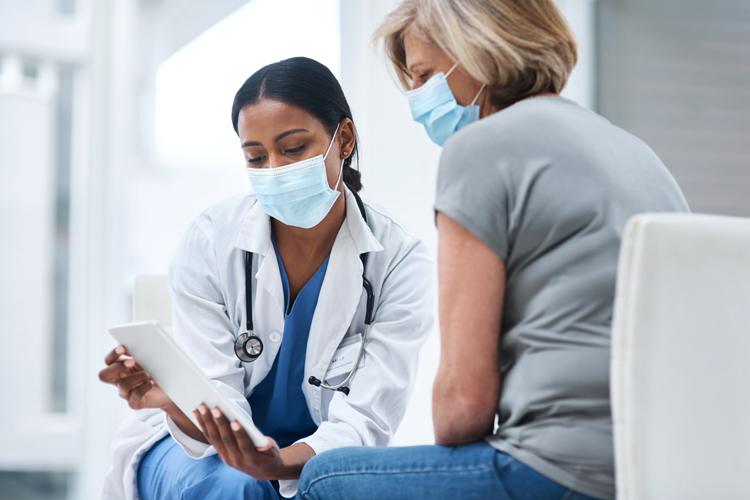 a woman in a white lab coat and surgical mask shows a table to a woman in a surgical mask sitting on the right