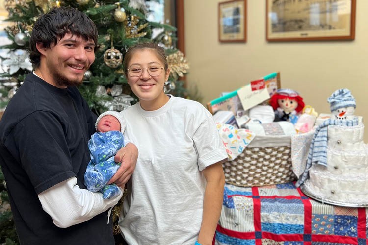 a man holding a newborn baby stands next to a woman, both smiling, a Christmas tree and a table with a basket of baby gifts are in the background