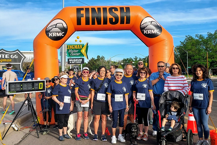 a group of people stand under an inflatable orange arch with the word "finish" printed on it. Signs in the background read "route 66 kingman" and "kingman visitor center"