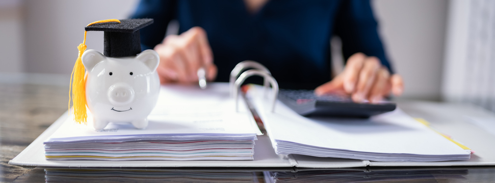 a piggy bank with graduation cap sits atop a binder of papers, a person's torso is in the background with a calculator