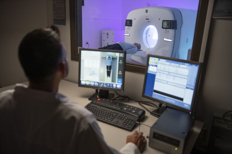 a medical professional looks through a window at a patient on a PET scanner while monitoring on screens