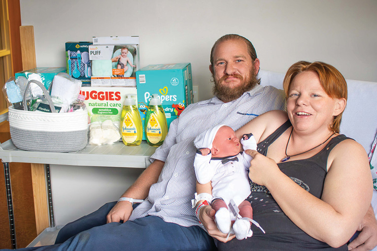 a couple holds their newborn baby in a hospital bed. next to them is a stand with a gift basket and baby supplies