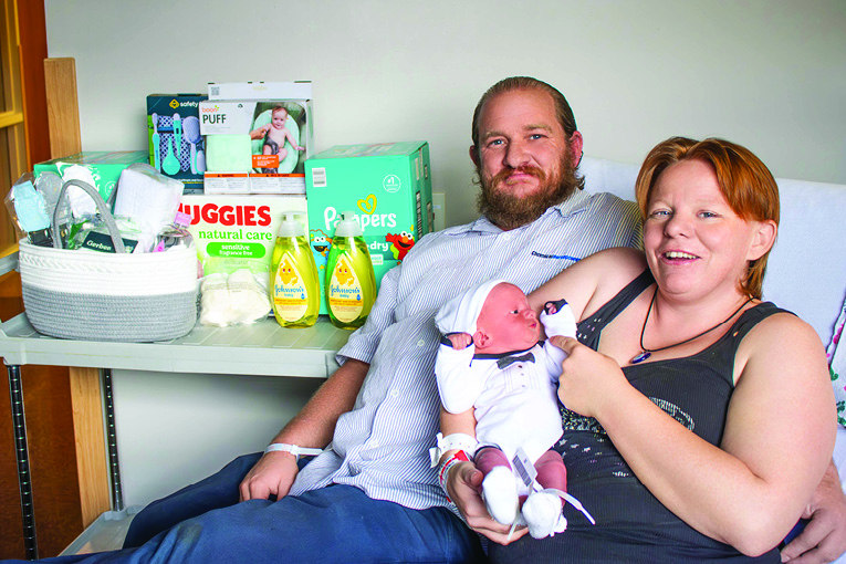 a couple holds their newborn baby in a hospital bed. next to them is a stand with a gift basket and baby supplies