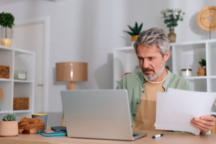 a man sitting at a desk looks at his laptop with papers in his hand