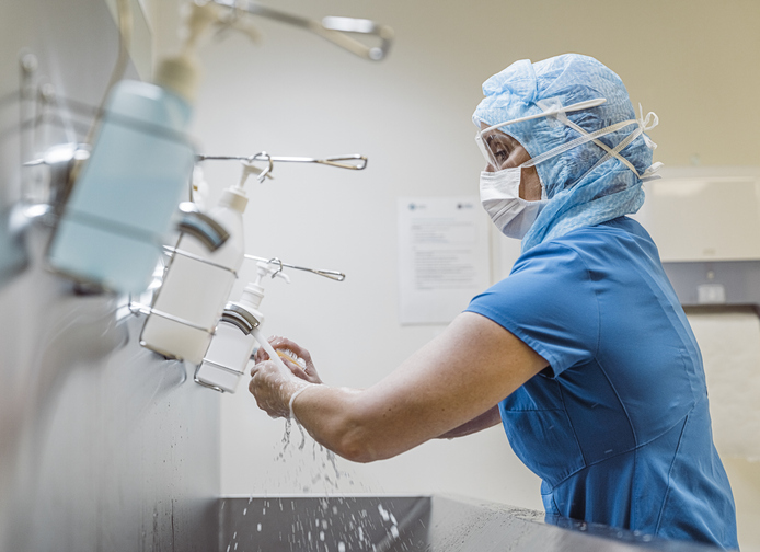 a woman in scrubs and PPE washes her hands at a metal sink