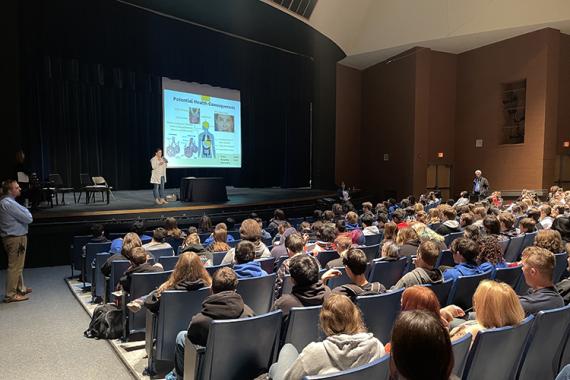 high school students sit in an auditorium facing a stage. Onstage is a presenter with a projection screen