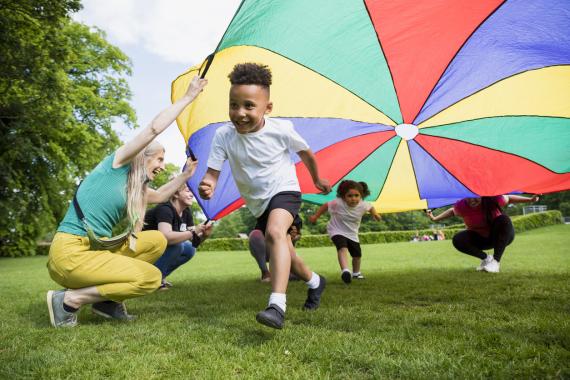 adults hold the edges of a colorful parachute while children run under it smiling