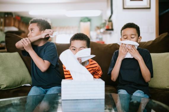 Three boys are sick at home, coughing and sneezing into tissues. They rest on the couch while they recover from their illness.